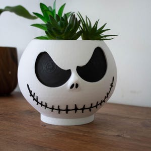Op de afbeelding: Een witte, bolvormige plantenbak met een Jack Skellington gezichtsontwerp, met zwarte oogkassen en een gestikte mond. De plantenbak bevat groene vetplanten en staat op een houten oppervlak. De tekst "GRADID-3D" is zichtbaar.