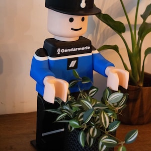 Peut inclure: Figurine imprimée en 3D représentant un gendarme français, de style bloc. La figurine porte un chapeau noir, un uniforme bleu et tient une petite plante en pot. La base est noire et la figurine a un visage souriant.