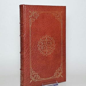 May include: A hardcover book with a textured, reddish-brown cover. The cover features a gold-colored, ornate border and a central medallion design. The spine is visible, and the book is propped up on a clear stand.