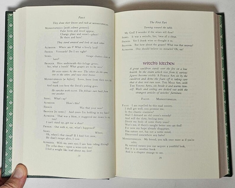 May include: Open book with text from a play, featuring pages with printed text in black ink. The book has a green cover with a decorative pattern and a hand is holding the book open.
