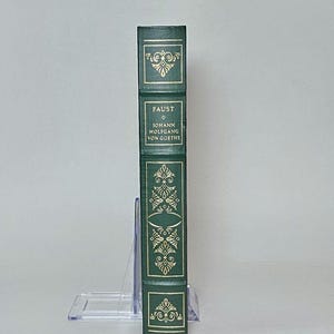 May include: A green hardcover book titled "Faust" by Johann Wolfgang von Goethe. The spine features gold decorative accents and the title in gold lettering. The book is displayed upright on a clear book stand.