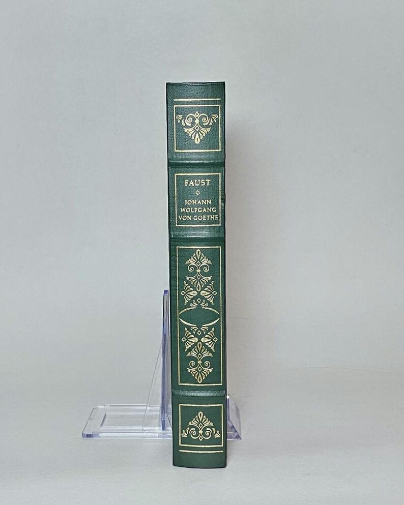 May include: A green hardcover book titled "Faust" by Johann Wolfgang von Goethe. The spine features gold decorative accents and the title in gold lettering. The book is displayed upright on a clear book stand.