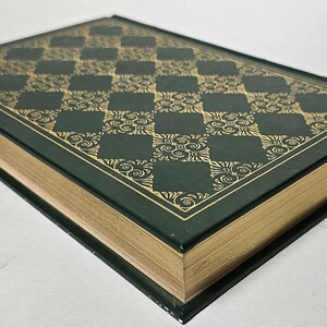 May include: A hardcover book with a dark green cover and gold-colored page edges. The cover features a gold checkered pattern with ornate floral designs. The book's spine is black, and the pages are gilded, giving it a classic, elegant look.