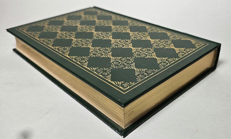 May include: A hardcover book with a dark green cover and gold-colored page edges. The cover features a gold checkered pattern with ornate floral designs. The book's spine is black, and the pages are gilded, giving it a classic, elegant look.