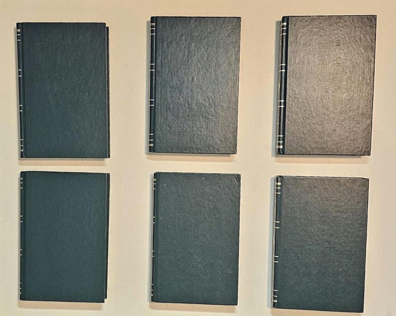 May include: Six hardcover books with dark teal covers are arranged on a light background. The spines have vertical silver accents. The books are rectangular and appear to be of similar size and style.