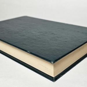 May include: A closed book with a dark teal cover and cream-colored pages. The book's cover has a textured appearance, and the edges are black. The pages are slightly yellowed, suggesting age or use. The book is lying flat.