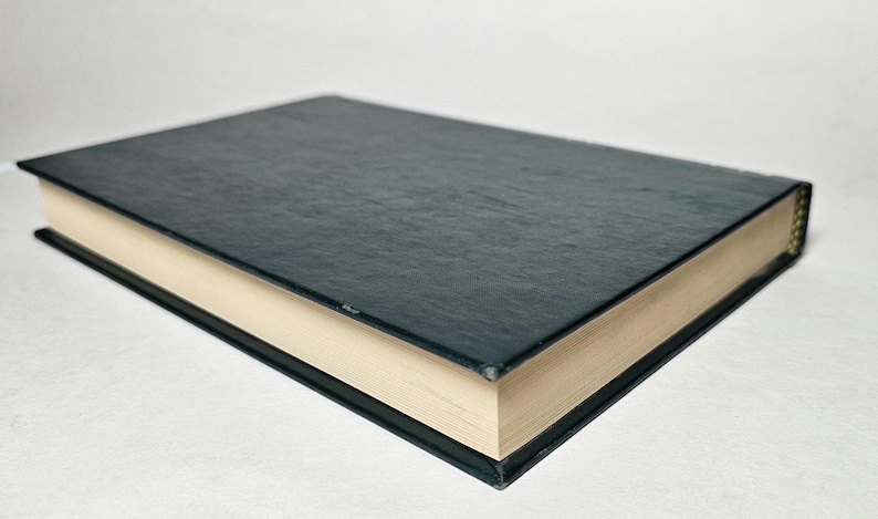 May include: A closed book with a dark teal cover and cream-colored pages. The book's cover has a textured appearance, and the edges are black. The pages are slightly yellowed, suggesting age or use. The book is lying flat.