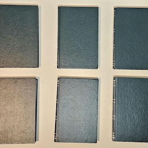 May include: Six hardcover books with dark green and gray covers are arranged in two rows. The spines have silver accents. The books are closed and appear to be in good condition. The covers are plain and without any visible titles.