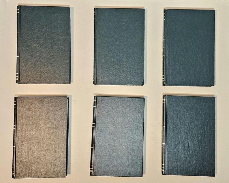 May include: Six hardcover books with dark green and gray covers are arranged in two rows. The spines have silver accents. The books are closed and appear to be in good condition. The covers are plain and without any visible titles.