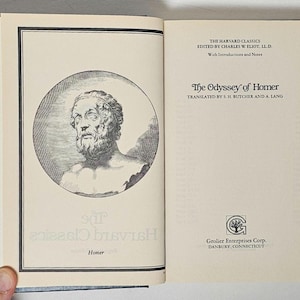 May include: Open book with black cover featuring a bust of a man in a circle on the left page. The right page displays the title "The Odyssey of Homer" and the text "The Harvard Classics" and "Grolier Enterprises Corp."
