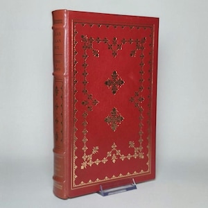 May include: A red hardcover book with gold foil detailing on the front cover and spine. The front cover features a decorative border and central floral-like patterns. The spine includes text that reads Edgar Allan Poe, Poems, and Essays.