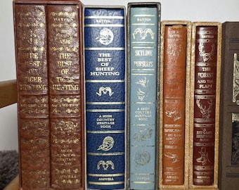 May include: A collection of six hardcover books with slipcases, featuring titles such as The Best of Tiger Hunting, The Best of Sheep Hunting, Skyline Pursuits, and The Forest and the Plain. The spines are in brown, blue, and tan with gold lettering.