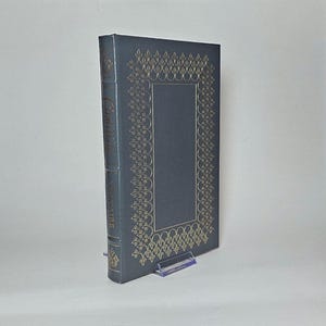 May include: A hardcover book with a dark blue cover and gold decorative accents. The book's spine displays the title "Candide" and the author's name. The cover features a gold border design around a rectangular space.