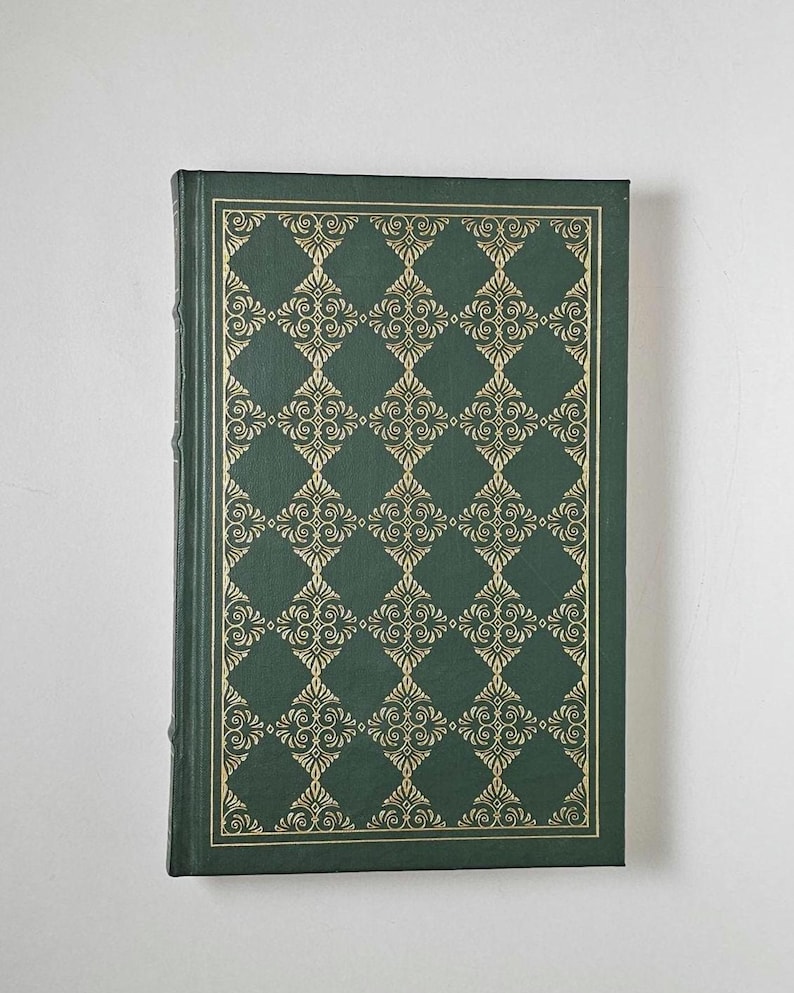 May include: A hardcover book with a dark green cover featuring a gold, ornate diamond pattern. The edges of the cover are also trimmed in gold. The book is closed and appears to be in good condition.