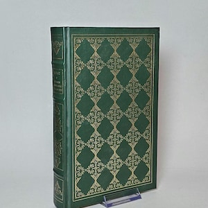 May include: A hardcover book with a green cover and gold decorative pattern. The spine and edges are also green. The book is propped up on a clear plastic stand. The title and author are visible on the spine.