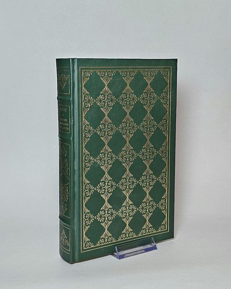 May include: A hardcover book with a green cover and gold decorative pattern. The spine and edges are also green. The book is propped up on a clear plastic stand. The title and author are visible on the spine.