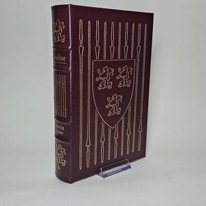 May include: A burgundy hardcover book titled "Ivanhoe" by Walter Scott. The cover features gold embossed illustrations of spears and a shield with lions. The spine has gold lettering and decorative elements.