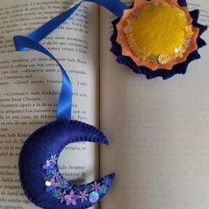Puede incluir: Dos marcadores de libro de fieltro, uno con forma de sol y el otro con forma de luna creciente. El marcador de sol es amarillo con fieltro naranja alrededor de los bordes y tiene pequeñas lentejuelas de colores. El marcador de luna es azul con lentejuelas moradas y azules. Ambos marcadores están en un libro con los números de página 194 y 195 visibles.
