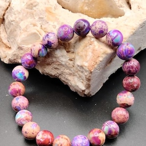 Puede incluir: Un brazalete de piedras preciosas de color púrpura y marrón con cuentas redondas. El brazalete está hecho de piedras naturales y tiene un patrón único de mármol.