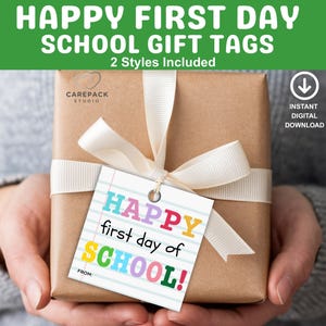 Peut inclure: Un cadeau emballé avec un ruban blanc et une étiquette indiquant "HAPPY first day of SCHOOL!" en lettres colorées. Le cadeau est enveloppé dans du papier brun et tenu par une personne.