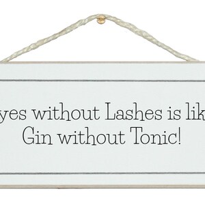 May include: A white wooden sign with black text that reads "Eyes without Lashes is like Gin without Tonic!" The sign is hanging from a white string with a gold tack.