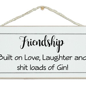 Può includere: Un cartello di legno bianco con testo nero che recita "Friendship Built on Love, Laughter and shit loads of Gin!" Il cartello ha un gancio di corda.