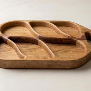 May include: Wooden serving tray with three compartments and integrated handles. The tray is made of light-coloured wood with darker wood accents. The compartments have curved edges and are designed for serving snacks or appetizers.