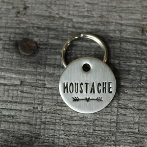 May include: Silver key chain with a round tag that says "Moustache" with an arrow and heart design.