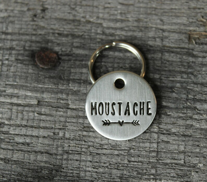 May include: Silver key chain with a round tag that says "Moustache" with an arrow and heart design.