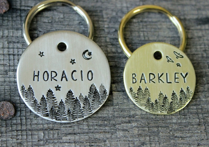 May include: Two silver and gold dog tags with engraved names "HORACIO" and "BARKLEY". Both tags feature a forest scene with trees and a crescent moon on the silver tag and birds on the gold tag.