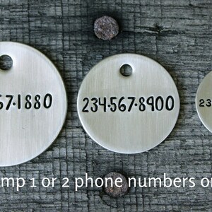 May include: Three silver round tags with phone numbers stamped on them. The numbers are 234-567-1880, 234-567-8900, and 234-567-8900. The text "I can stamp 1 or 2 phone numbers on all tags." is below the tags.
