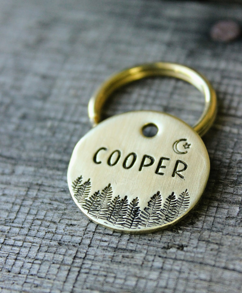 May include: A gold-coloured metal dog tag with the name "COOPER" engraved on it. The tag features a crescent moon and star design and a silhouette of a forest with ferns.