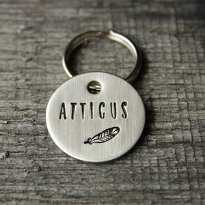 May include: Silver dog tag with a feather design and the name "Atticus" engraved on it.