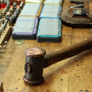 May include: A close-up of a hammer with a wooden handle and a copper-coloured head. The hammer is lying on a wooden workbench with various tools and supplies in the background.