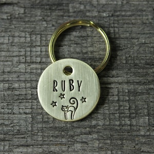 May include: A gold-colored metal pet tag with the name "RUBY" engraved on it. The tag also features a small cat silhouette and three stars.