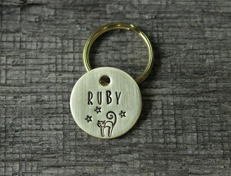 May include: A gold-colored metal pet tag with the name "RUBY" engraved on it. The tag also features a small cat silhouette and three stars.