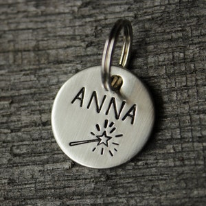 May include: Silver dog tag with the name "ANNA" engraved on it. A small star with a line extending from it is engraved below the name.