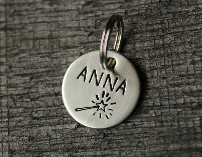 May include: Silver dog tag with the name "ANNA" engraved on it. A small star with a line extending from it is engraved below the name.