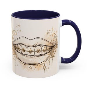 Puede incluir: Taza de cerámica blanca con interior y asa azul marino. La taza presenta una ilustración detallada de una boca sonriente con brackets y adornos dorados. El diseño incluye acentos de flor de lis y elementos decorativos.