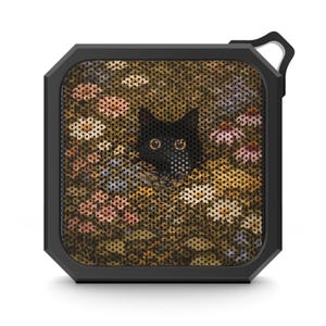 May include: Black square portable speaker with a black cat peeking out from a field of flowers. The speaker has a black frame and a small loop for hanging. The floral design includes pink, white, and yellow flowers.