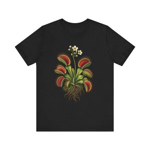 May include: A black short-sleeved t-shirt featuring a central graphic illustration of a Venus flytrap plant. The plant has green leaves with red interior traps, spiky edges, visible brown roots, and a tall stem topped with small white flowers.