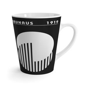 Puede incluir: Taza de café con leche de estilo Bauhaus en blanco y negro. La taza presenta la palabra "BAUHAUS" y el año "1919" en texto blanco. Un diseño geométrico blanco está en la parte delantera de la taza. La taza tiene un asa blanca.
