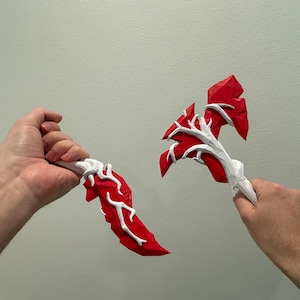 May include: Two hand-carved wooden weapons with a red and white paint scheme. The weapons are shaped like a dagger and an axe. The axe has a white handle and a red blade with white veins. The dagger has a red blade with white veins and a white handle.