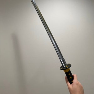 May include: A silver katana sword with a black and gold handle. The sword is held by a hand.
