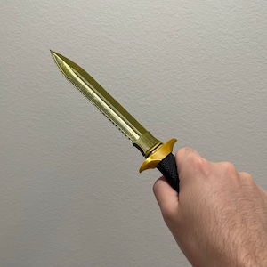 May include: A gold-colored dagger with a black handle. The blade has a serrated edge.