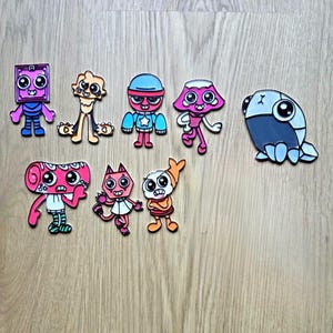 May include: A collection of colorful cartoon character stickers. The stickers feature various designs, including a square-shaped character, a cat-like figure, and a creature with a mushroom-shaped head. The stickers are in shades of pink, blue, orange, and purple.