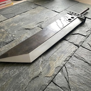 May include: A large, heavy-duty sword featuring a wide, flat blade with a dark, weathered metallic finish and a light-colored, beveled edge. The hilt area includes a rectangular guard with circular cutouts and a long, cylindrical handle grip.