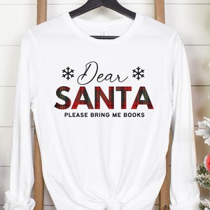 Puede incluir: Una camiseta blanca de manga larga con el texto negro Dear y la palabra SANTA en un patrón de cuadros rojo y verde, flanqueada por dos iconos de copos de nieve negros. Debajo, el texto dice PLEASE BRING ME BOOKS en una fuente negra simple.