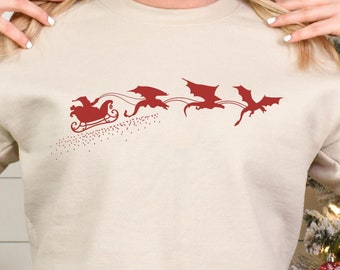 May include: Sand-colored sweatshirt featuring a red graphic of a sleigh pulled by dragons. The design includes a sleigh with a driver and three dragons in flight. The word "Sand" is in a white label in the bottom right corner.
