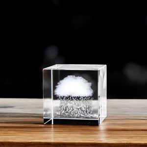 Può includere: Un blocco di vetro trasparente e quadrato con un disegno di nuvola bianca e pioggia all'interno. La nuvola è soffice e la pioggia appare come piccole goccioline trasparenti. Il blocco è posto su una superficie di legno.